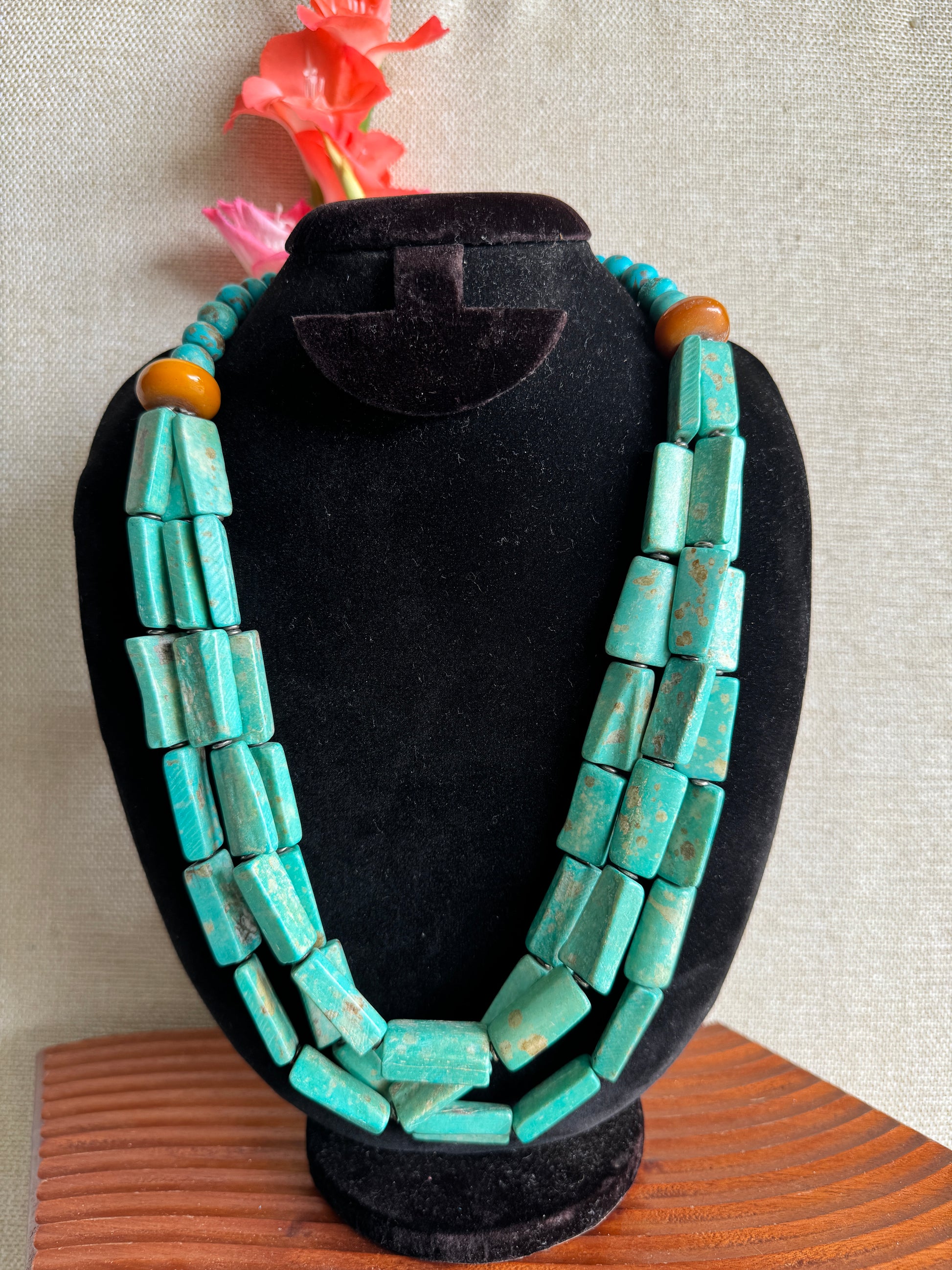 Turquoise beaded bracelet on a black velvet stand with a pink flower and wooden surface in the background.
